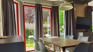 A dining area with a table, chairs, a television, and a view of a garden through large windows. - Wildgans 48 - Klassisches Skandinavisches Ferienhaus für bis zu 6 Personen (Mirow)