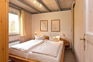 Una habitación con dos camas, vigas de madera, un ventanal con cortinas, luces fijadas a la pared y cuadros enmarcados.