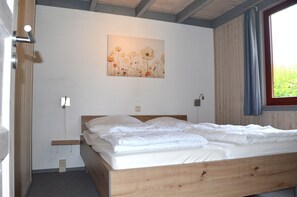 Ein Schlafzimmer mit einem hölzernen Bett, einer weißen Wand, einem Blumenbild und einem Fenster mit Vorhängen.