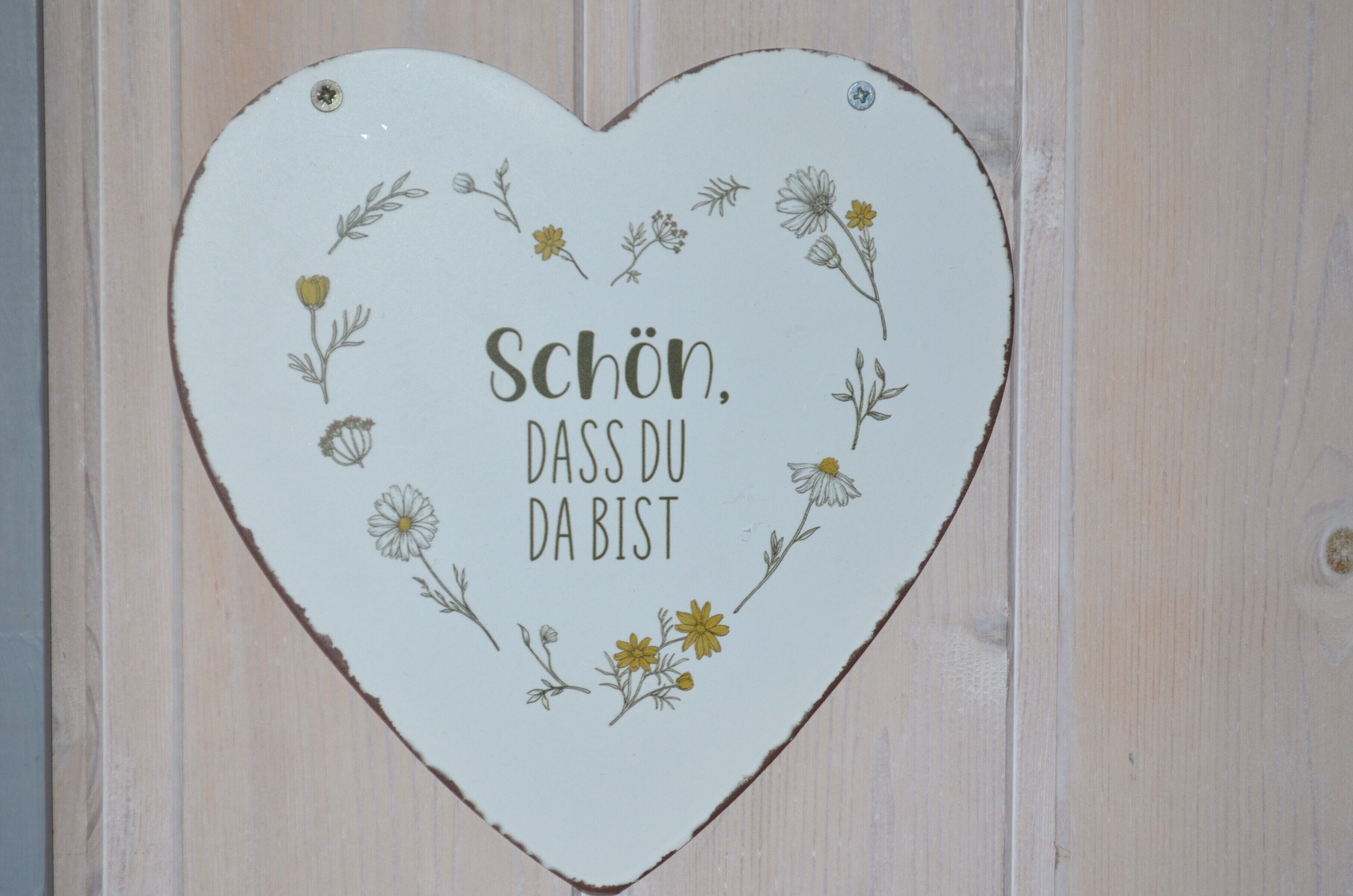 Un letrero con forma de corazón, adornado con motivos florales y el texto 'Schön, dass du da bist' en una pared de madera.