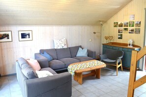 Ein gemütliches Wohnzimmer mit einer Couch, einem kleinen Tisch und einem Sessel. An den Wänden hängen Bilder in Rahmen und es steht eine Holzkommode bereit.