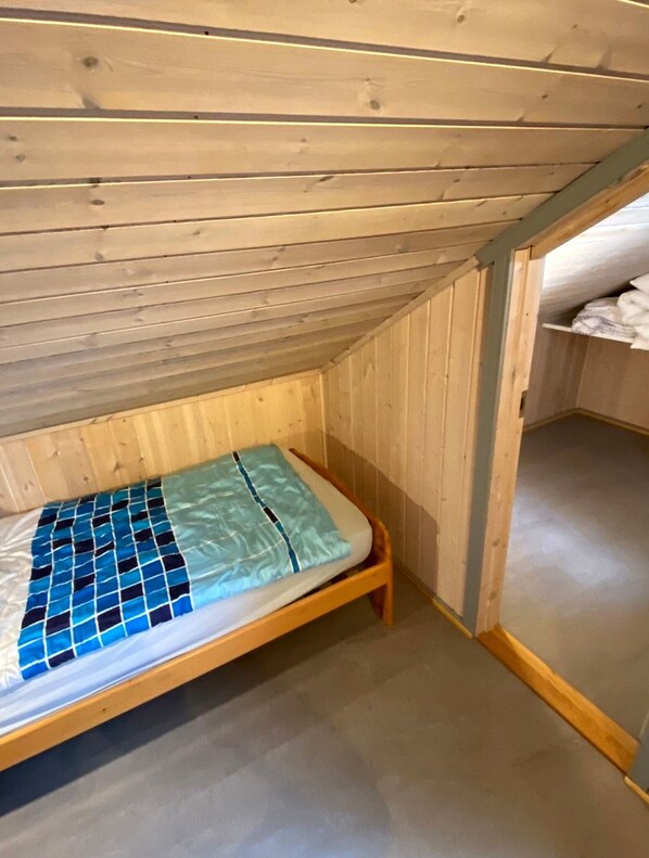 Une petite chambre mansardée avec un plafond en pente en bois, un lit simple recouvert d’une courtepointe à motifs bleus et blancs, et un banc en bois.
