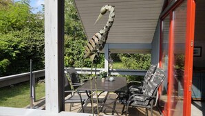 Eine Terrasse im Freien mit Tisch und Stühlen, einer dekorativen Vogel-Skulptur und einer Glastür, die in einen Garten führt.