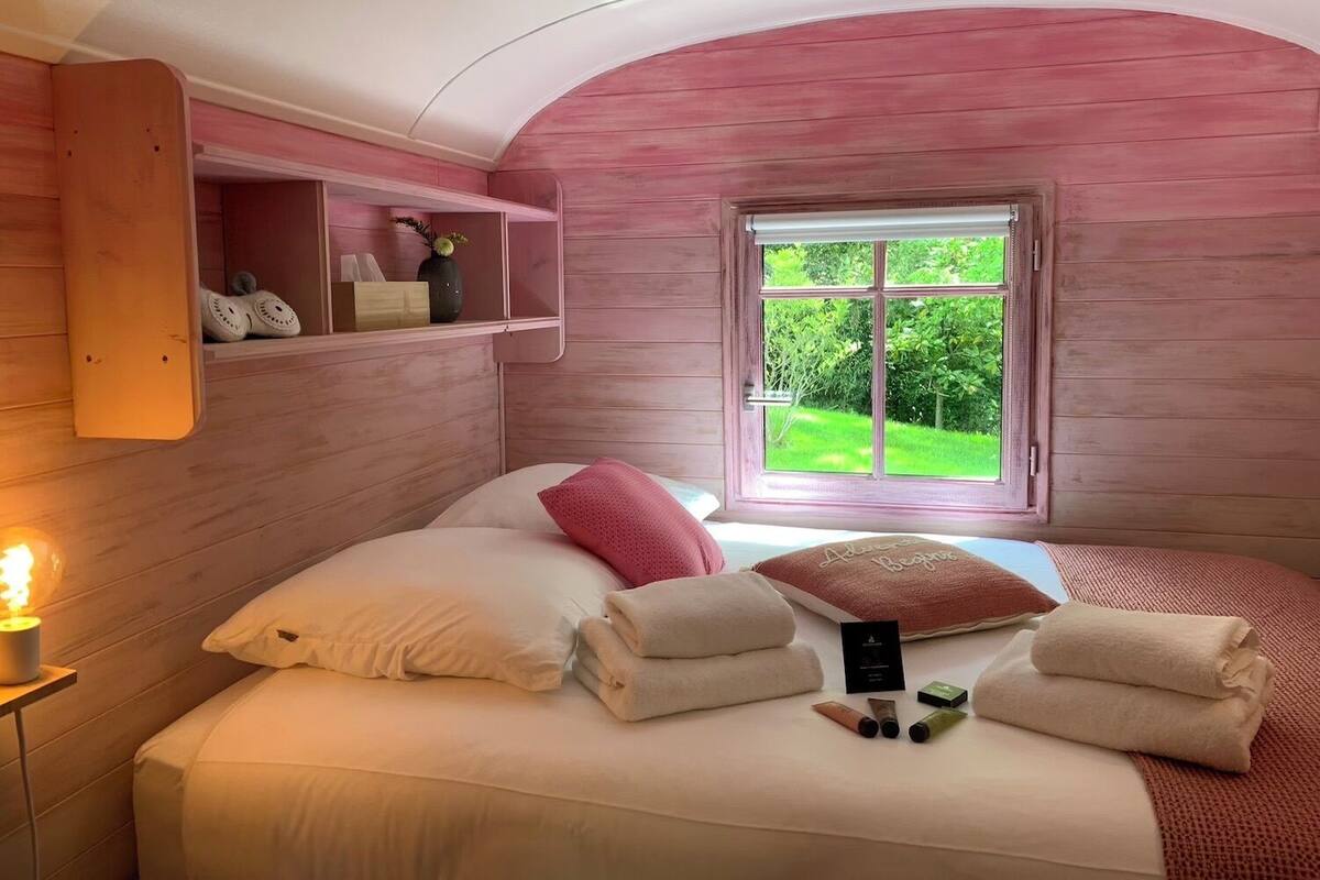 Ein gemütliches Schlafzimmer mit Bett, Kissen, Handtüchern, einem Nachttisch mit Lampe, einer Ablage für Toilettenartikel und einem Fenster mit Blick ins Grüne.