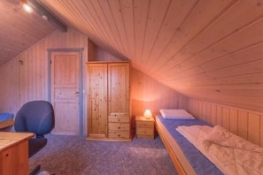 Una habitación pequeña y acogedora con paredes de madera y techo inclinado. Cuenta con una cama, un escritorio, una silla y un armario.