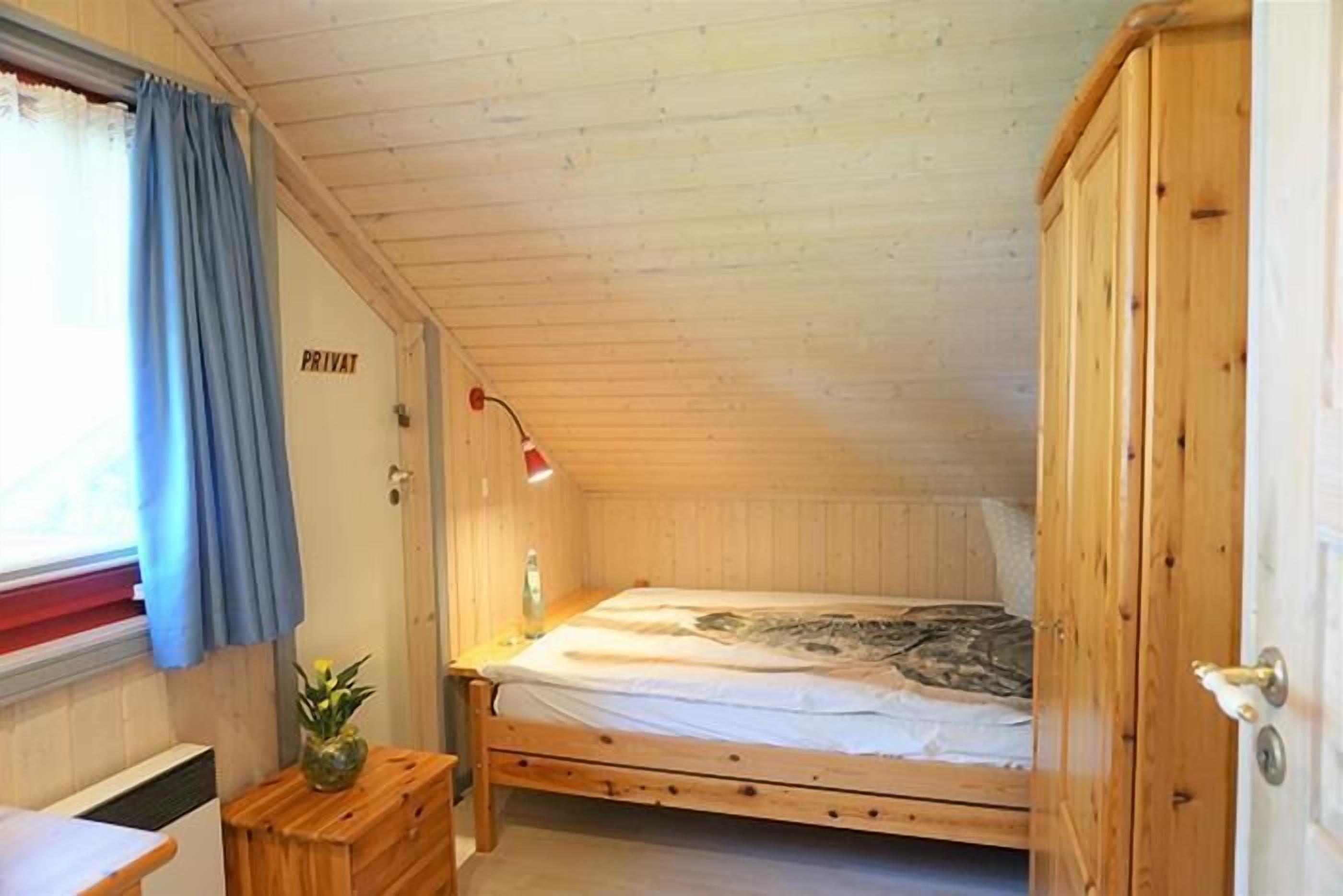 Quarto pequeno com painéis de madeira, uma cama de solteiro, mesa de cabeceira com abajur e janela com cortinas.