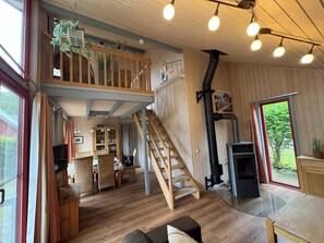 Interior de madera con entrepiso, escalera, chimenea y ventanas amplias.