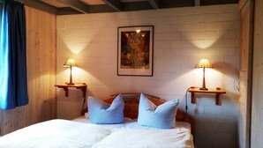 A bedroom with a wooden ceiling, a bed with white bedding, two blue pillows, two bedside tables with lamps, and a framed artwork on the wall.