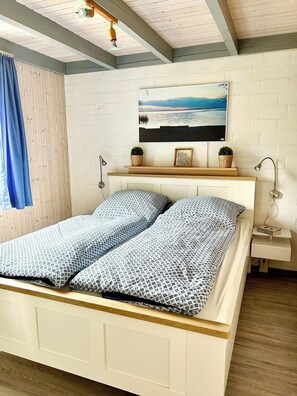 A bed with a patterned bedspread, a bedside table with a lamp, a wall-mounted shelf with decorative items, and a framed picture above the bed. - Wildgans 63 - Klassisches Skandinavisches Ferienhaus für bis zu 6 Personen (Mirow)