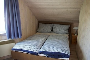 Ein kleiner, gemütlicher Raum mit Holzboden und -wänden, einem Einzelbett mit gemusterten Betttextilien, einem Nachttisch mit Lampe und einem Fenster mit Vorhängen.
