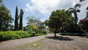 Pátio pavimentado com uma árvore, um edifício ao fundo e vegetação ao redor.