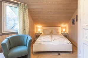 Cozy single-room accommodation with a sloped ceiling, featuring a neatly made bed, bedside tables, and a comfortable armchair.