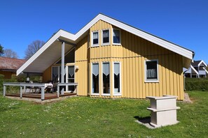 A yellow house with a sloped roof, a porch with chairs, and a small garden with flowers. - Wildgans 13A - Klassisches Skandinavisches Ferienhaus für bis zu 6 Personen (Mirow)