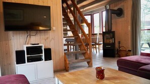 A cozy room with a wooden interior, a flat-screen TV mounted on the wall, a wooden staircase, a fireplace, and a table with a colorful vase. - Wildgans 113 - Klassisches Skandinavisches Ferienhaus für bis zu 6 Personen (Mirow)