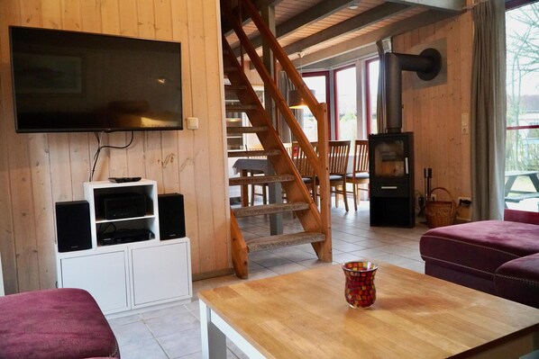 A cozy room with a wooden interior, a flat-screen TV mounted on the wall, a wooden staircase, a fireplace, and a table with a colorful vase.