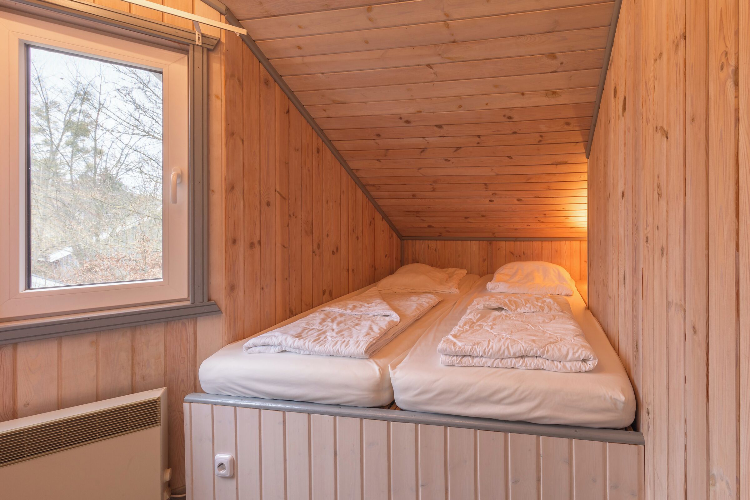 A small, wooden cabin room with two beds, a window, and a radiator.