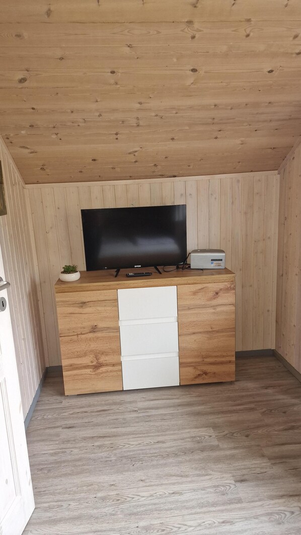 A small, cozy room with a wooden ceiling and walls, a flat-screen TV on a wooden stand, a small plant, and a white drawer unit.