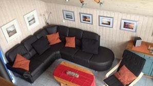 Un salon avec un canapé d’angle noir, un pouf rouge et deux fauteuils en bois.