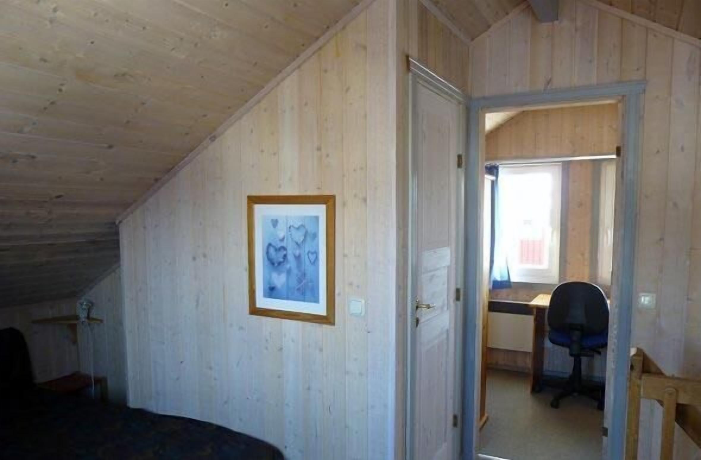 Et værelse med trævægge, et indrammet billede, et skrivebord med stol, og en dør til et andet rum.