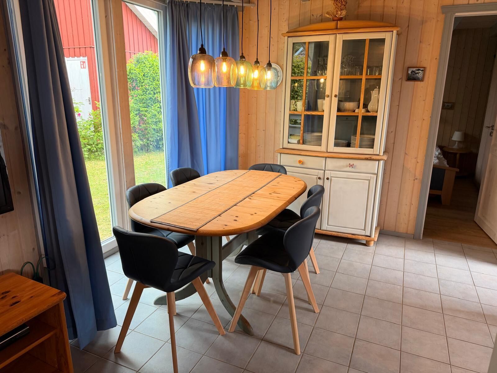 A dining area with a wooden table and chairs, a cabinet with glass doors, and a view of greenery outside.
