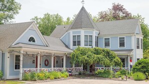 Une grande maison à deux étages, dotée d’une véranda qui fait le tour de la maison, d’une petite structure en forme de tour et d’un jardin bien entretenu.