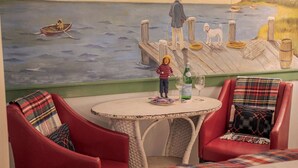Une pièce chaleureuse ornée d’une fresque représentant un quai et des bateaux, avec une table ronde sur laquelle se trouvent une bouteille et des verres, ainsi que des fauteuils rouges à l’assise rembourrée et dotés de coussins à motifs.