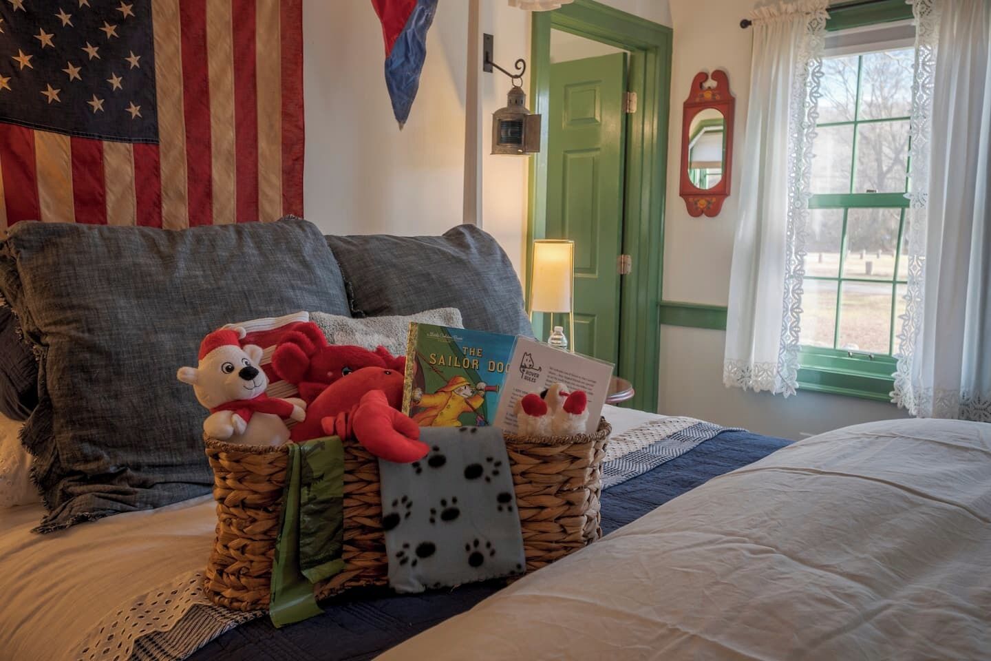 Een netjes opgemaakt bed met een decoratief kussen, een mand met knuffels en een boek, en een ingelijst certificaat op het nachtkastje.