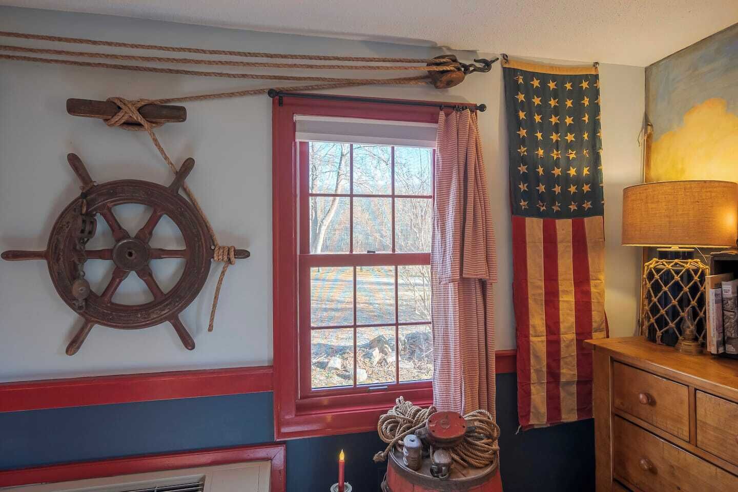 Ett rum med ett träskeppsratt, ett fönster med röd ram, en amerikansk flagga och en lampa på en träbyrå.