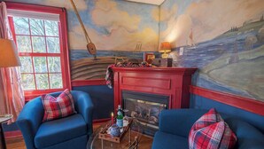 A cozy room with a fireplace, two blue armchairs, a small table with a bottle and glasses, and a mural of a seascape. - New England Hospitality at the Carriage House
Mystic Vacation Rentals LLC (Old Mystic)