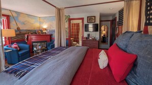 A hotel room with a large bed, a seating area with a blue armchair, a fireplace, a television, and American flags. - New England Hospitality at the Carriage House
Mystic Vacation Rentals LLC (Old Mystic)