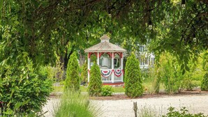 A gazebo with a white roof and red trim, surrounded by lush greenery and a gravel path. - New England Hospitality at the Carriage House
Mystic Vacation Rentals LLC (Old Mystic)