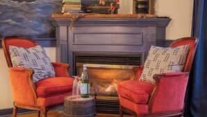 Una sala de estar acogedora con chimenea, dos sillones rojos tapizados con cojines estampados, una mesita con una botella y copas, y un cuadro con temática marina sobre la chimenea.