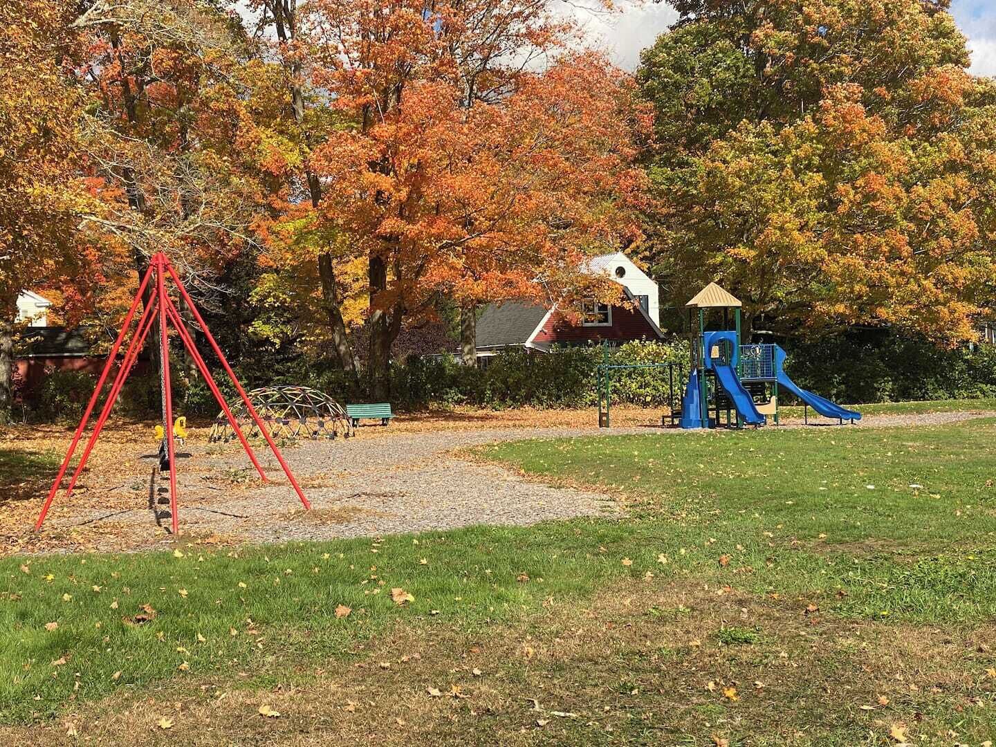 Un'area giochi con altalene rosse e uno scivolo blu, circondata da alberi con foglie autunnali.