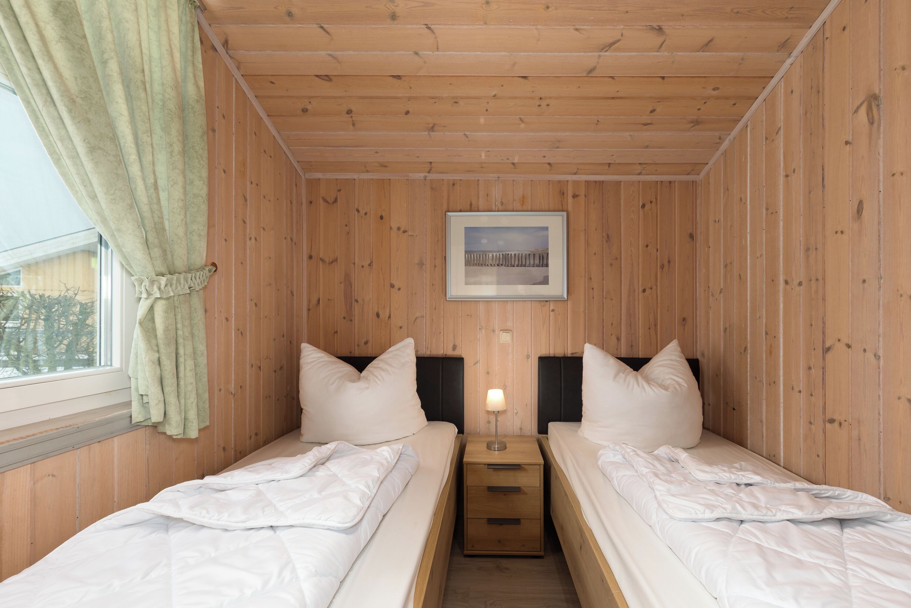 Una habitación con paneles de madera, dos camas con ropa blanca, una mesita de noche con una lámpara y un cuadro enmarcado en la pared.