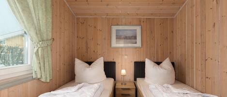 Una habitación con paneles de madera, dos camas con ropa blanca, una mesita de noche con una lámpara y un cuadro enmarcado en la pared.