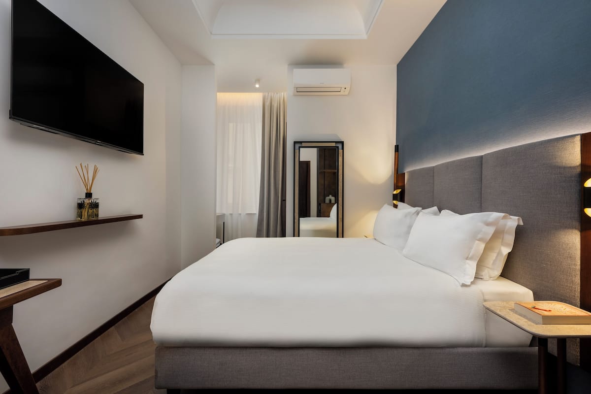 A modern hotel room with a large bed, a flat-screen TV mounted on the wall, a bedside table with a book, and a small shelf with decorative items.