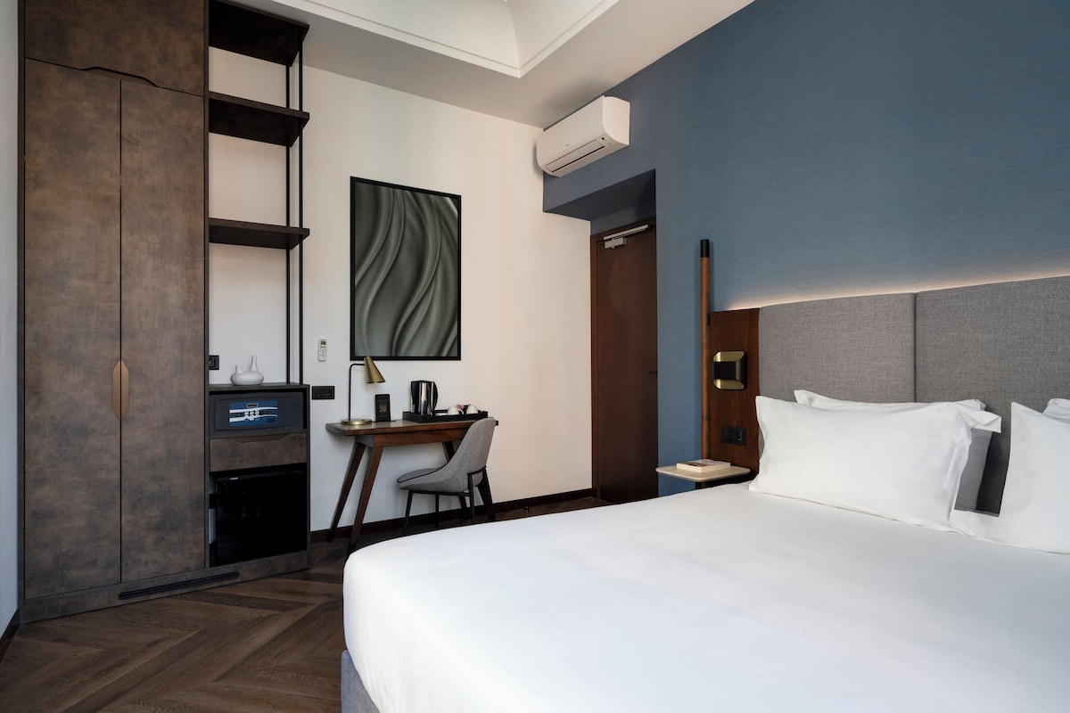 A modern hotel room with a large bed, a desk with a chair, a wardrobe, and an air conditioning unit.
