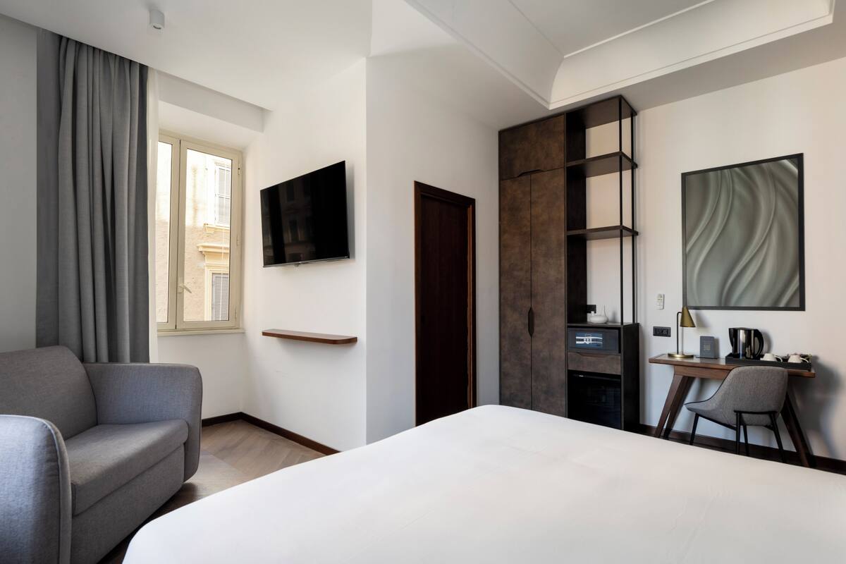 A modern hotel room with a large bed, a desk with a chair, a TV mounted on the wall, and a wardrobe.
