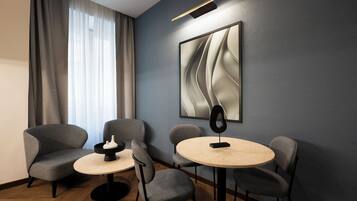 Una habitación moderna con una mesa redonda y cuatro sillas grises, un cuadro abstracto grande en la pared, y una ventana con cortinas traslúcidas.
