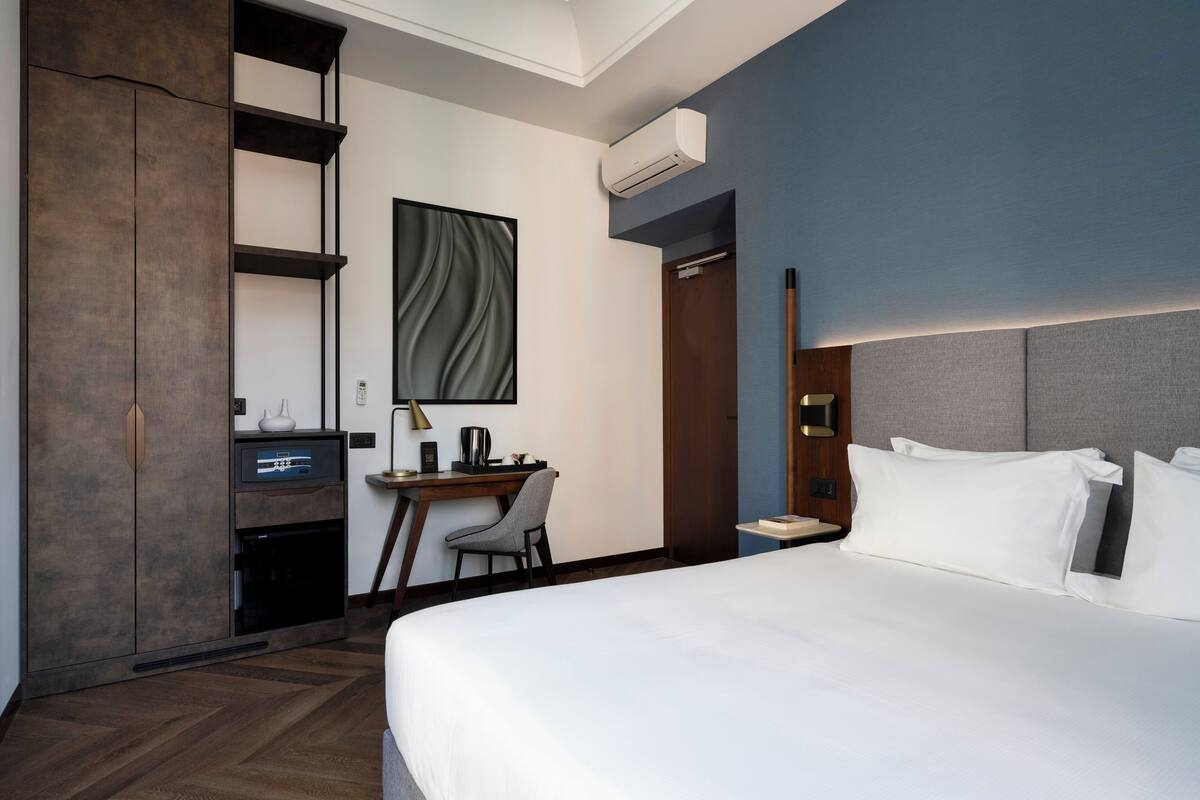 A modern hotel room with a large bed, a desk with a chair, a wardrobe, and an air conditioning unit.
