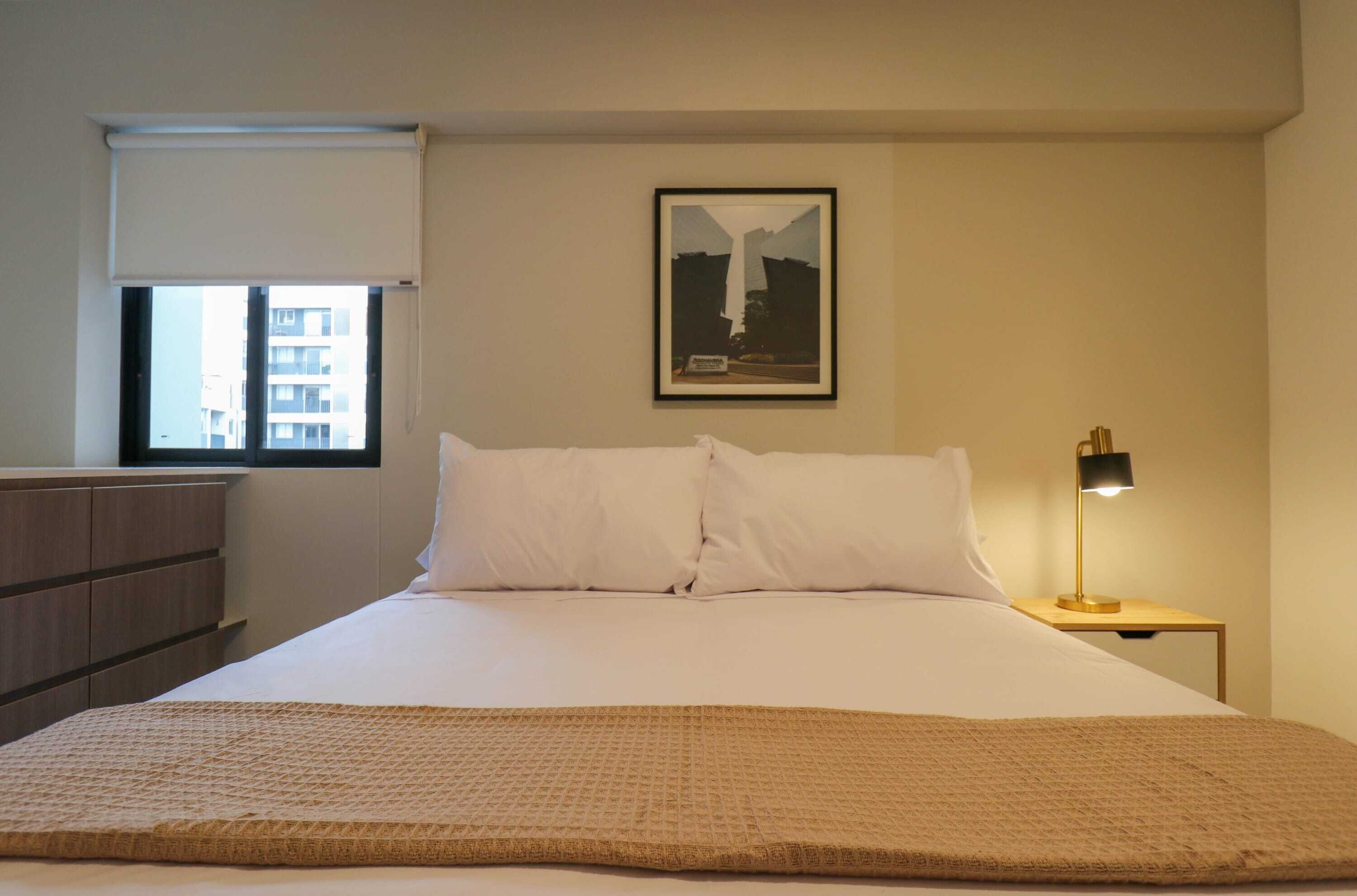 Camera d'albergo con un letto, un comodino con una lampada, una finestra vista edifici e un quadro appeso al muro.
