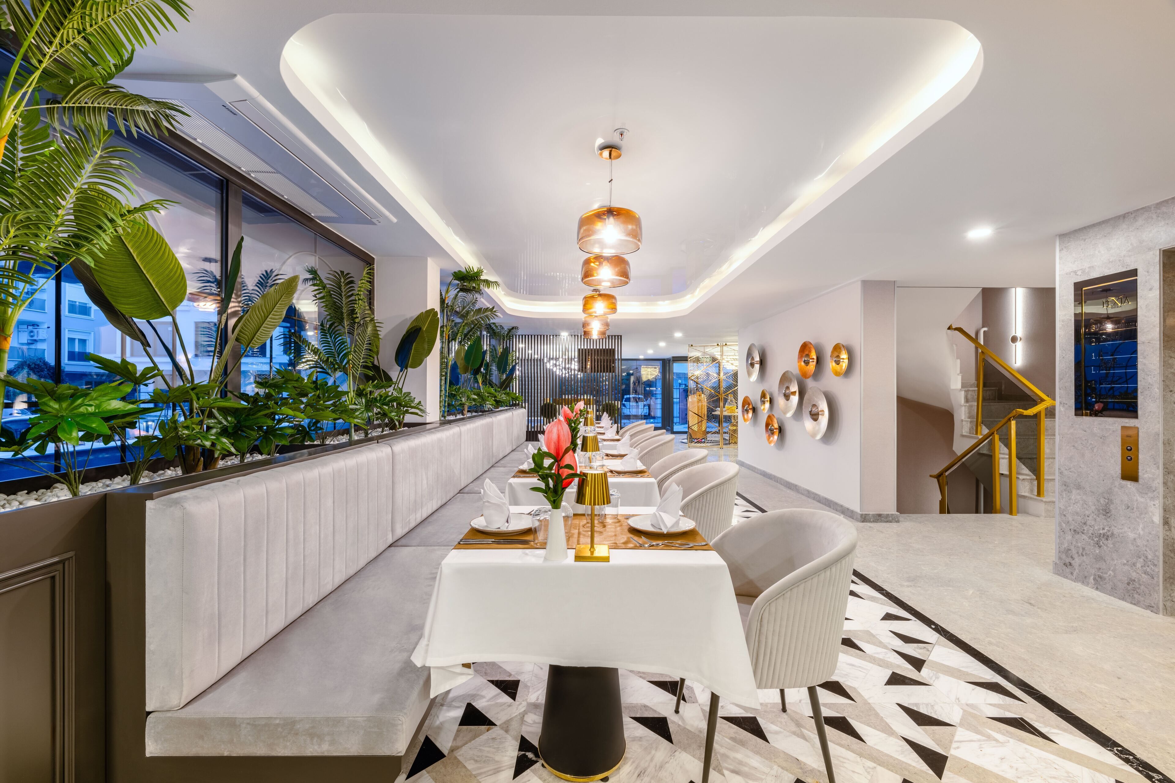 A modern dining area with a long table set for a meal, surrounded by plants and a view of the cityscape.