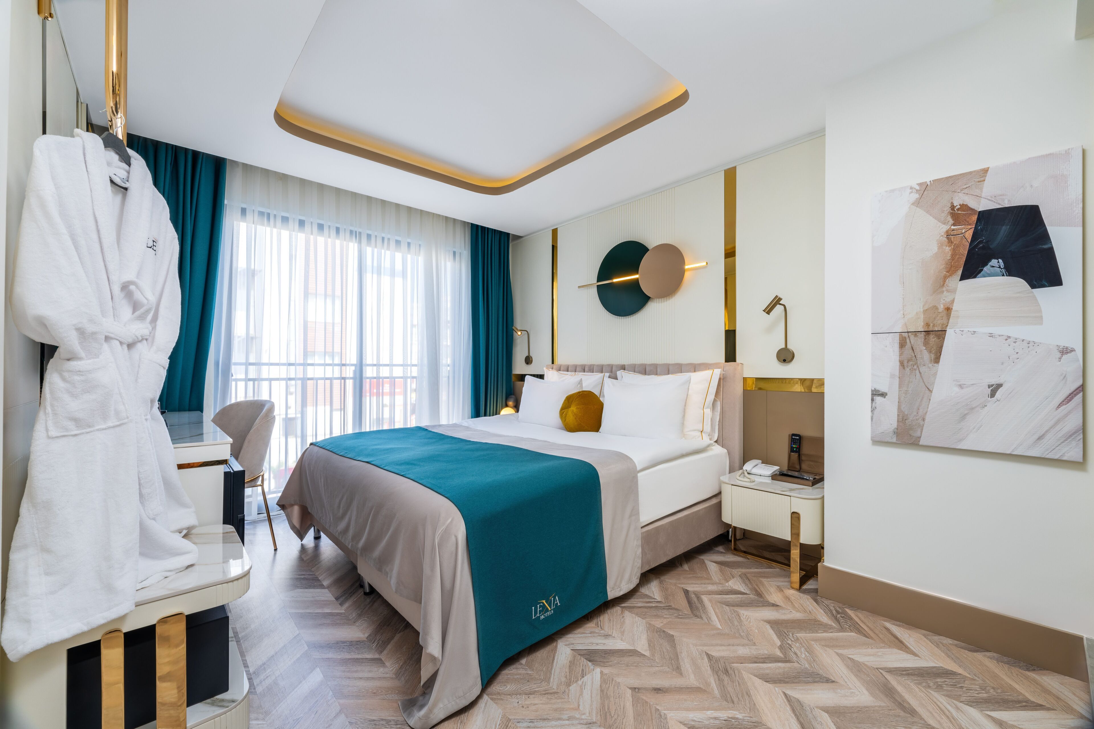 A hotel room with a bed, a desk, a chair, a robe, a window with curtains, a wall-mounted lamp, and a painting.