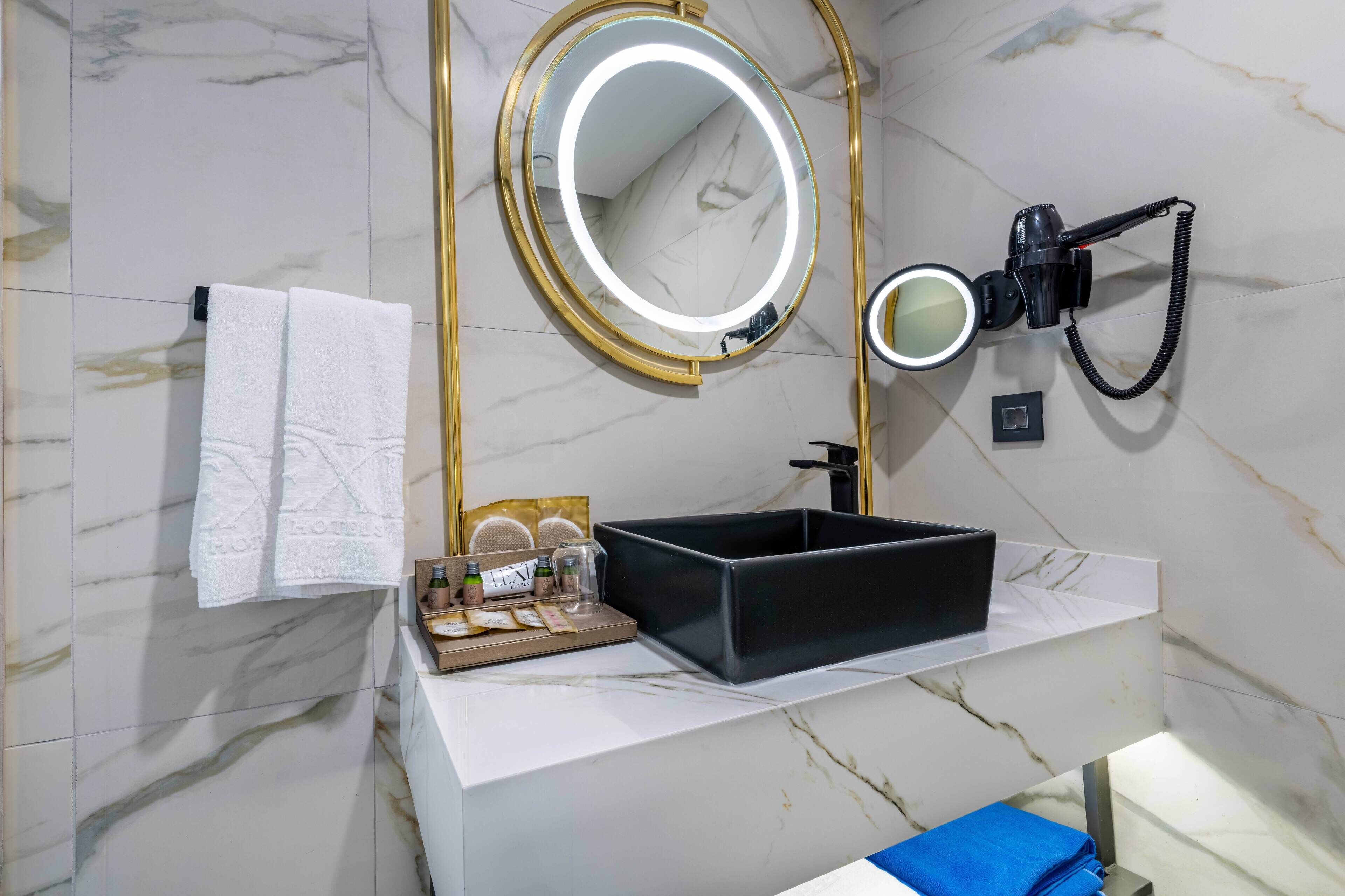 A bathroom with a marble-tiled wall, a white countertop, a black sink, a gold-framed mirror, and a hairdryer.