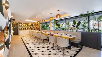 A modern restaurant with a geometric patterned floor, multiple dining tables with chairs, pendant lighting, and a view of greenery through large windows.