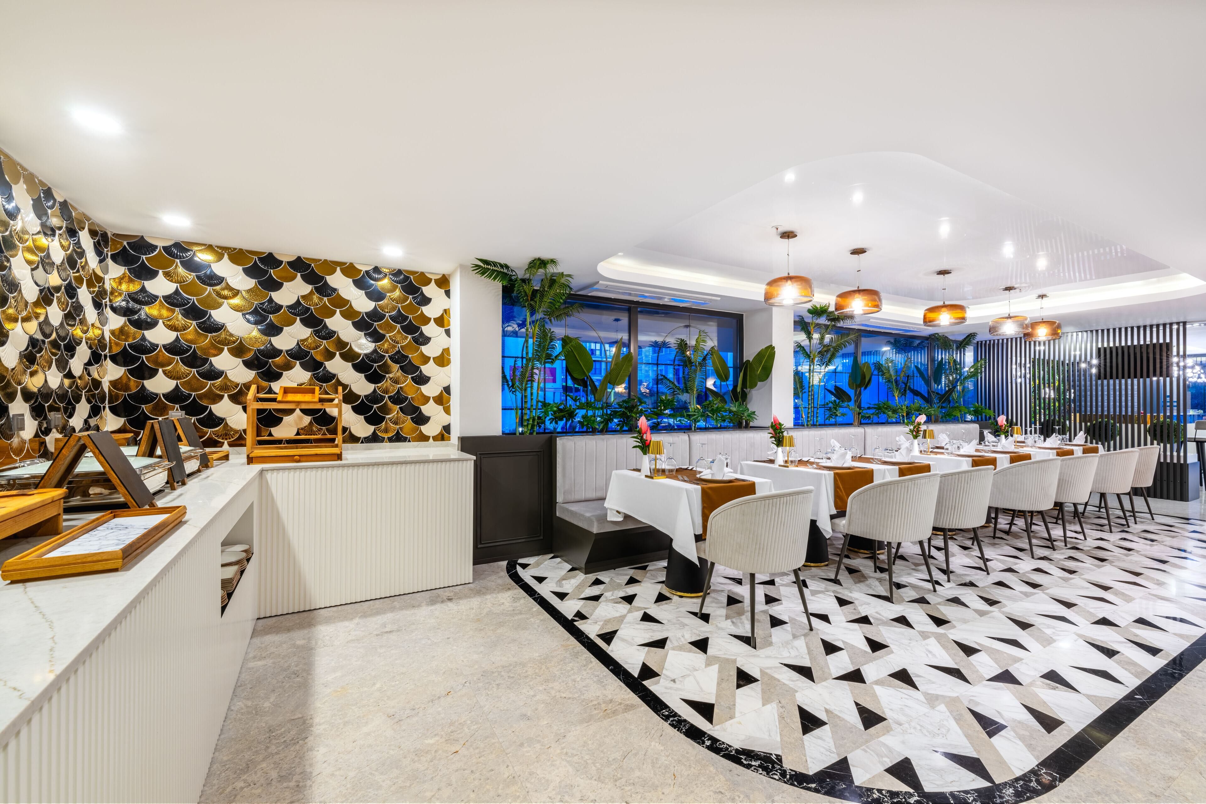 A modern restaurant with a white and black geometric floor pattern, a long counter with framed pictures, and a seating area with white chairs and a table set for a meal.