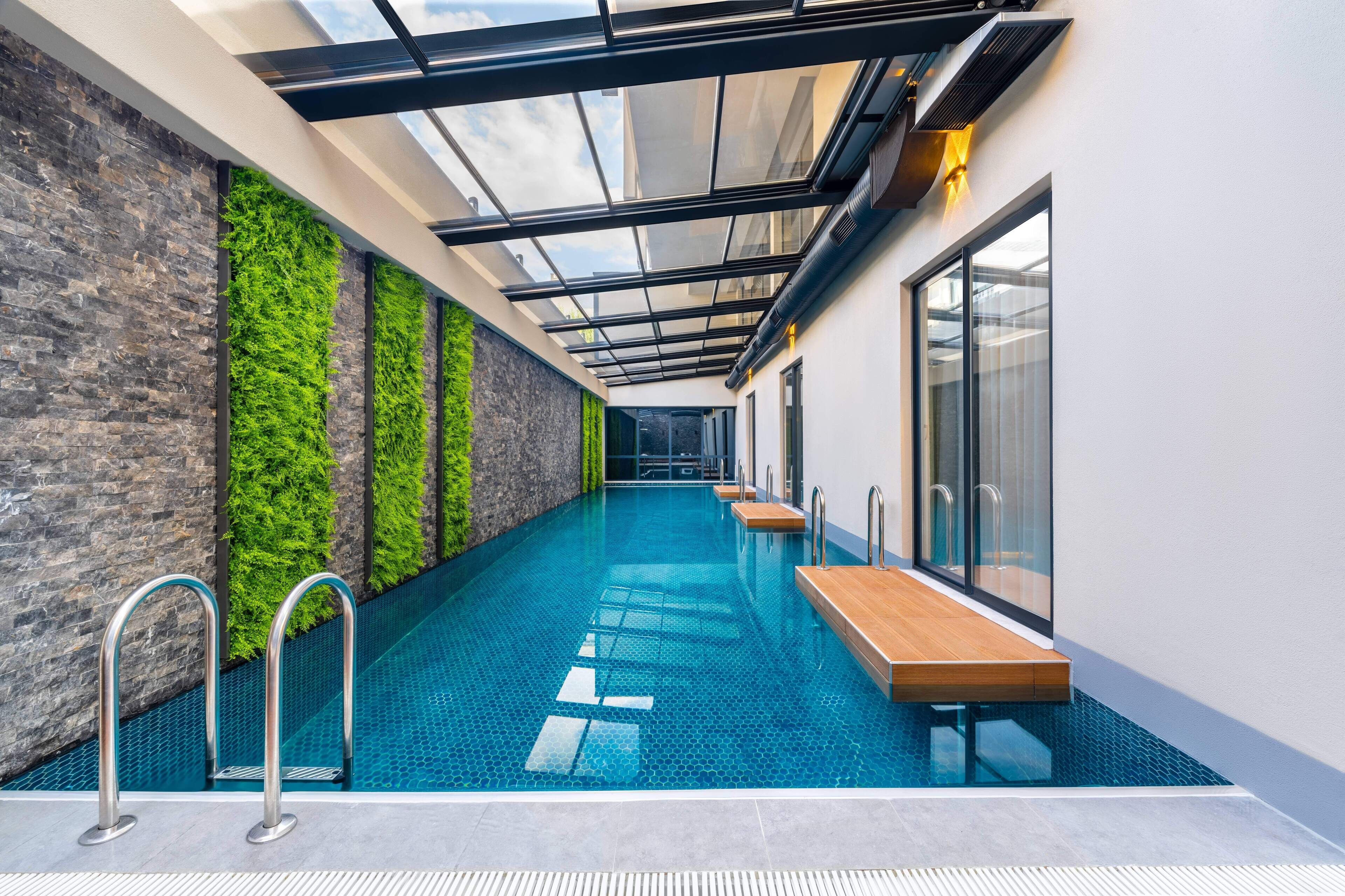 Ein moderner, überdachter Schwimmbereich mit Glaswänden, Holzterrasse und einer grünen Wand.