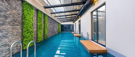 Una piscina interior moderna con paredes de vidrio, terraza de madera y una pared verde.