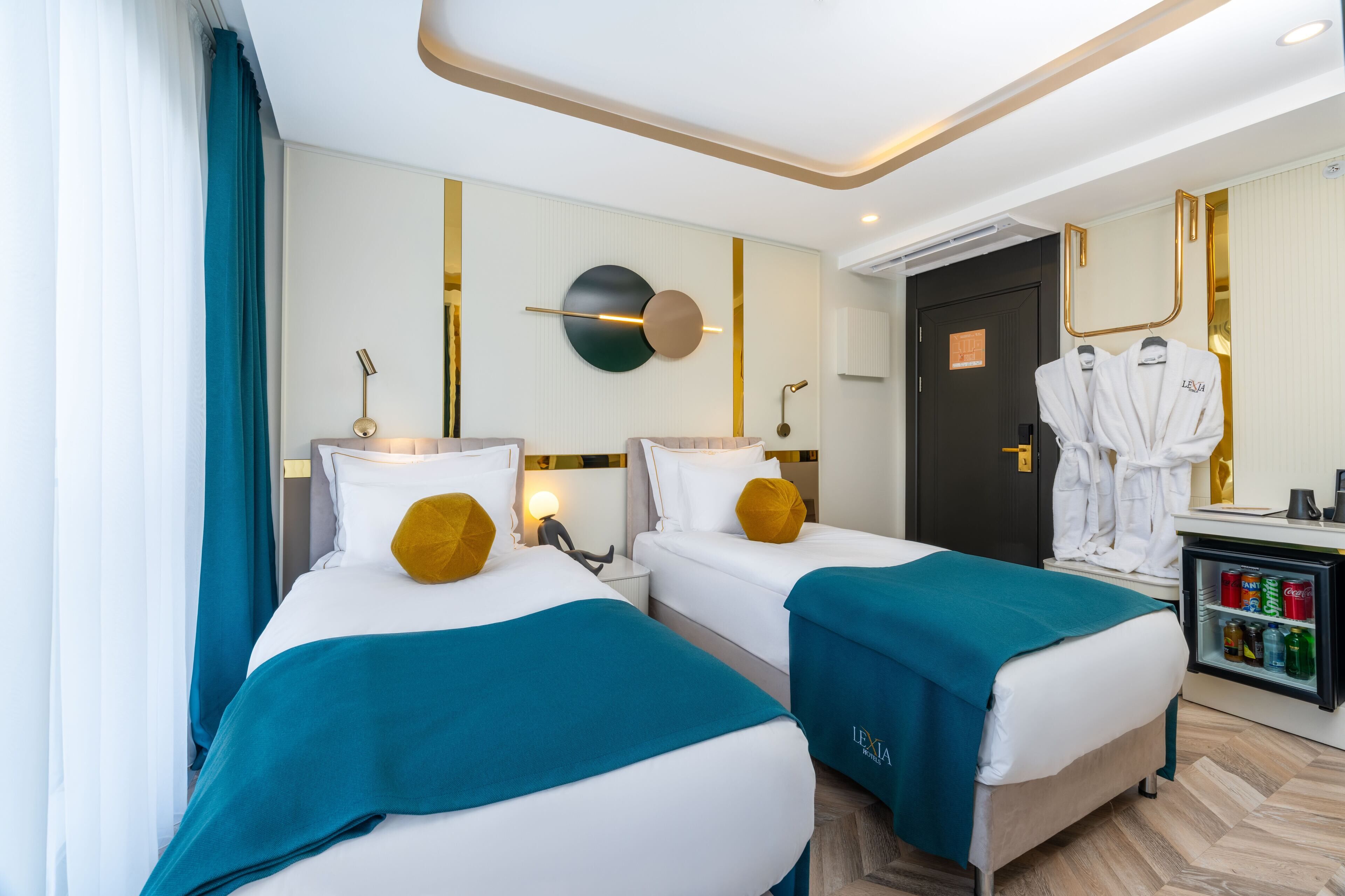 A hotel room with two beds, teal bedspreads, white bedding, a minibar, and a mirror with a gold frame.