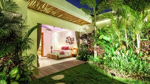 Un dormitorio moderno con piso de madera, una cama con almohadones rojos y una mesita de noche. Se observa un pequeño jardín con diversas plantas y una terraza de madera que conduce a la habitación.